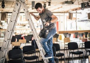 A male event planner climbs a ladder to hang stage lights.