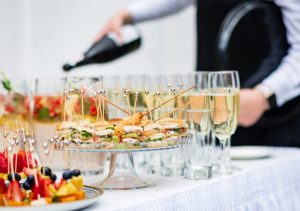 An event table has a white tablecloth and holds champagne glasses, appetizers, and fruit.