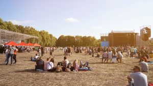 People sit in the grass at an outdoor festival venue with a stage.