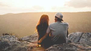 A man and woman sit on top of a mountain while watching the sun set.