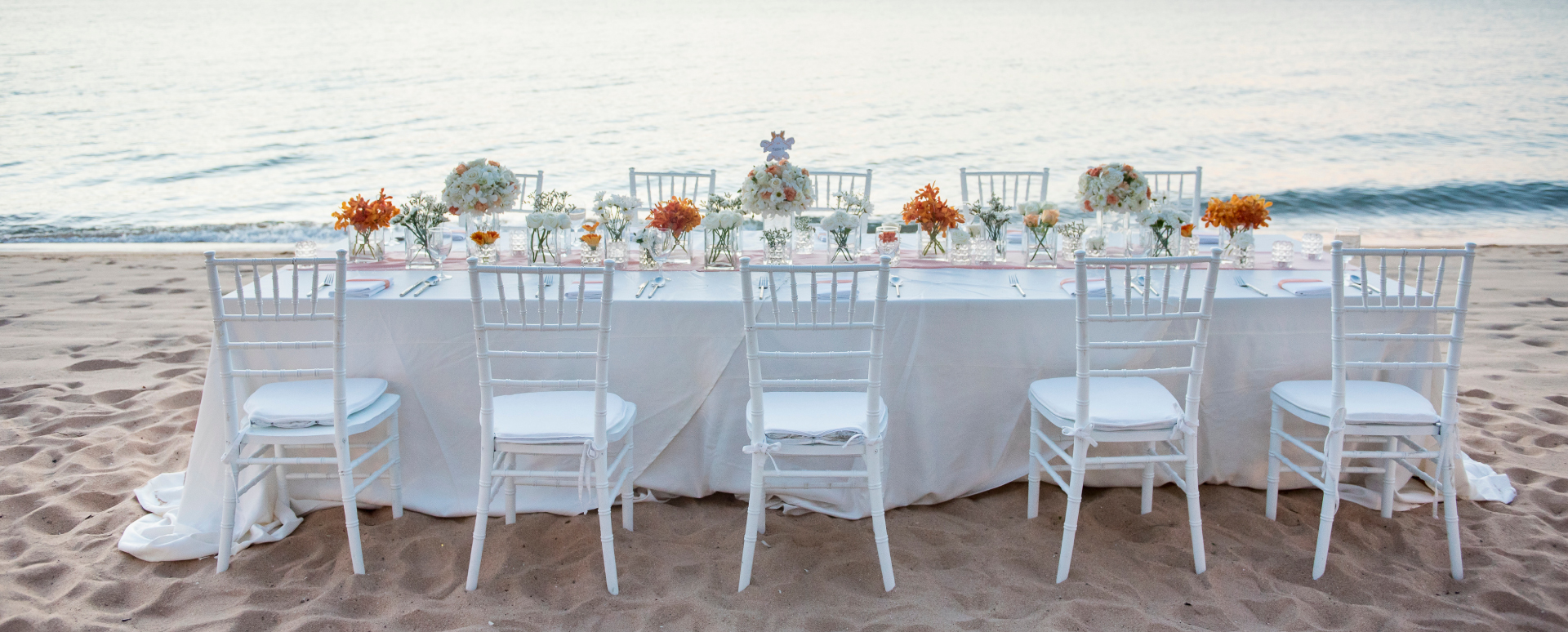 Table for a wedding with white tablecloth, 10 white chairs, and flowers in vases on a beach near the ocean.
