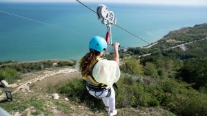 Woman in blue helmet zip lines down a mountain near the ocean.