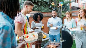 Six people gather around a BBQ smiling as they wait for the food to cook.