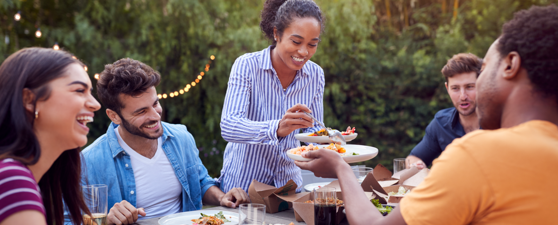 Five people of various ethnicities sit outside at a picnic table smiling and eating.