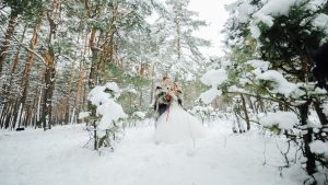 A couple stand in a snowy field as they get photos taken of them in their wedding attire.