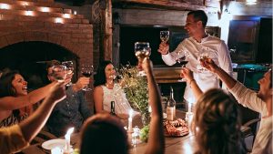 Six people enjoy a candlelit dinner at a bistro and toast the chef, who stands at the head of the table.