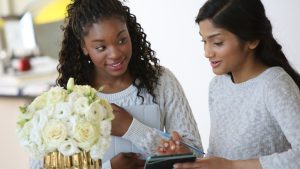 Two women in white sweaters review a list next to a wedding bouquet with cream colored flowers.