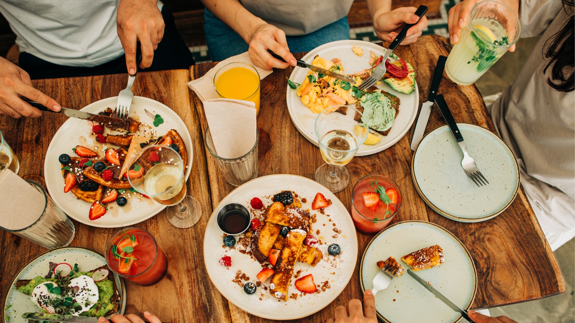 Five people sit at a restaurant table with plates of brunch items that include strawberry waffles, avocado toast, and scrambled eggs.