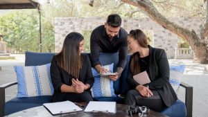 A man and two women gather near a blue couch in a courtyard reviewing their event planning notes on clipboards.