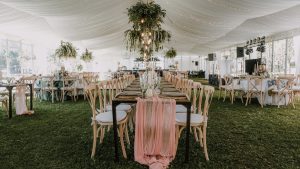 White wedding tent with plants and lighting hanging from the ceiling above long tables with wood chairs and centerpieces.