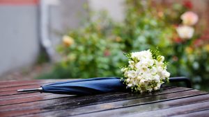A blue umbrella rests on a wooden surface, adorned with a small bouquet of white flowers, set against a blurred floral background.